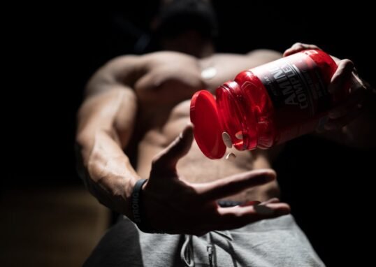 Shirtless muscular man pouring protein supplements from a bottle, showcasing fitness and strength.