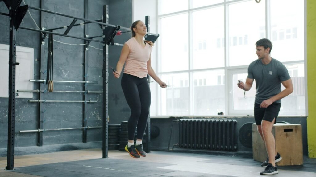 Mulher pulando corda na academia enquanto homem observa, exemplificando treino de cardio.